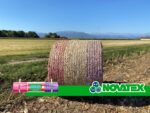 Rotoballa avvolta da rete bianca con strisce rosa e viola, fascia Novatex e rotolo etichettato Rekord ML 2600 in un campo soleggiato.